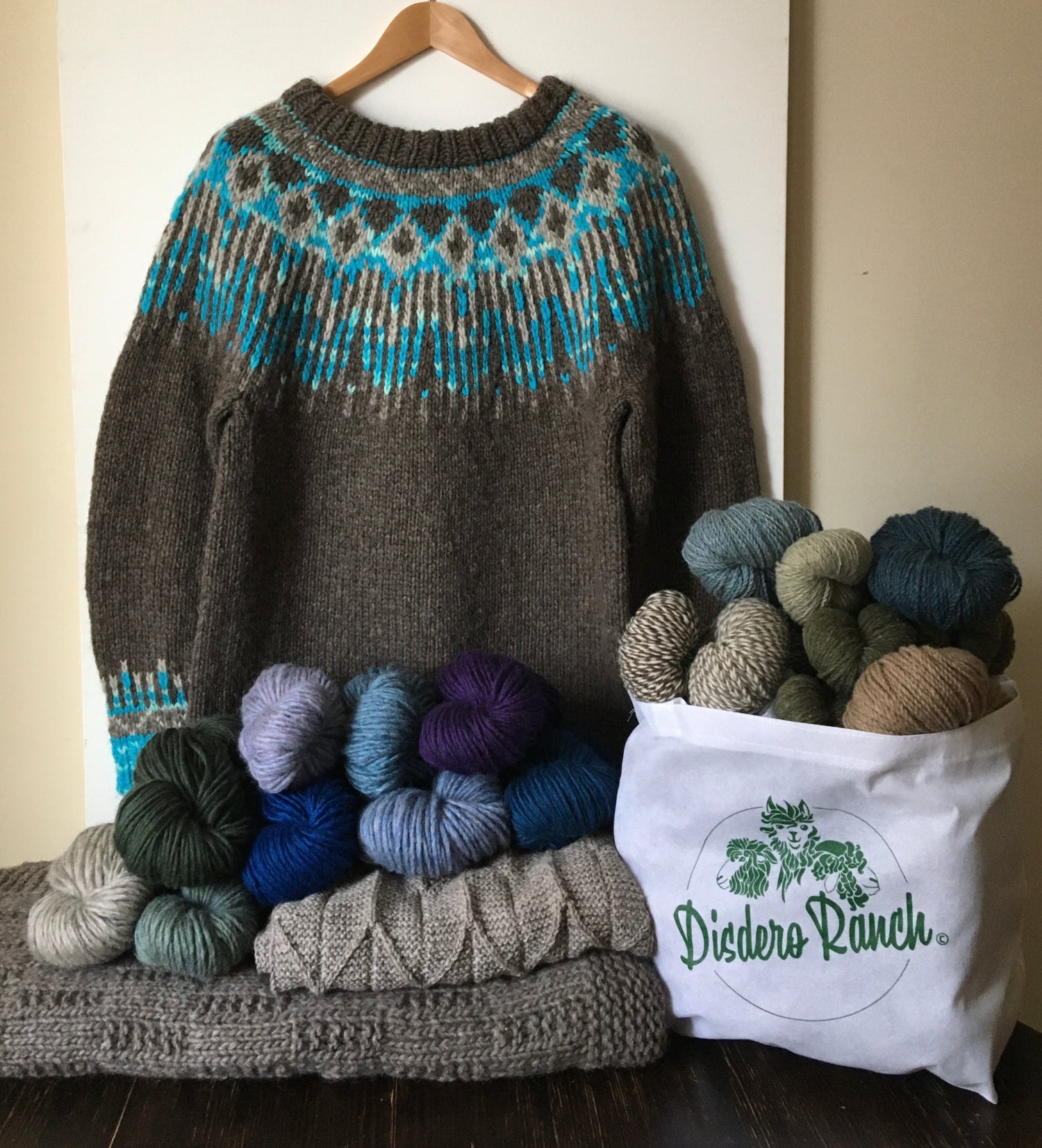 Display of knitted sweater, shawl, and blanket on table with coloured hanks of yarn